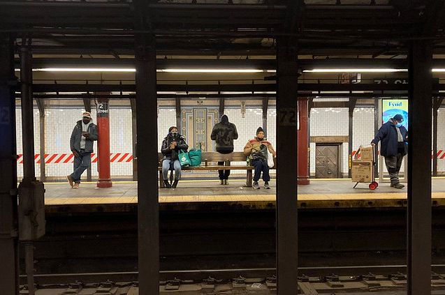 Several commuters spread out across a 72nd Street subway platform, framed by beams.